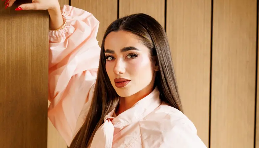 Lola Lolita, con su cabello largo y liso, posa con un vestido rosa pálido, apoyando su mano en un pilar de madera.