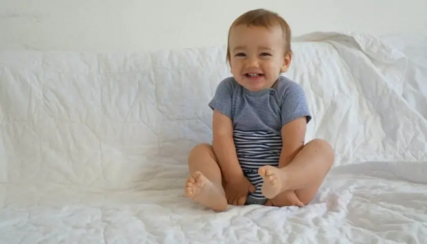 Lächelndes Baby im gestreiften Body sitzt auf weißer Decke.