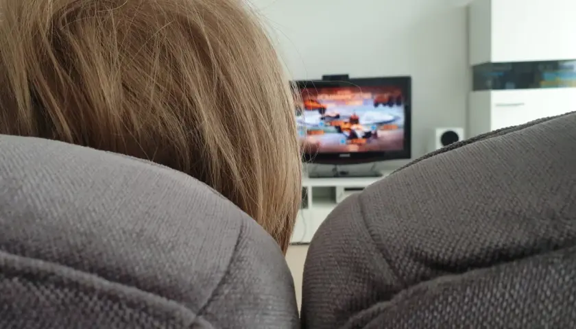 Niemowlę z blond włosami siedzi na kanapie, a w tle widać włączony telewizor z grą.