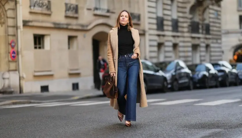 Une femme porte un jean flare bleu, idéal pour allonger la silhouette et sublimer toutes les morphologies. Elle est habillée d'un long manteau camel et d'un col roulé noir.