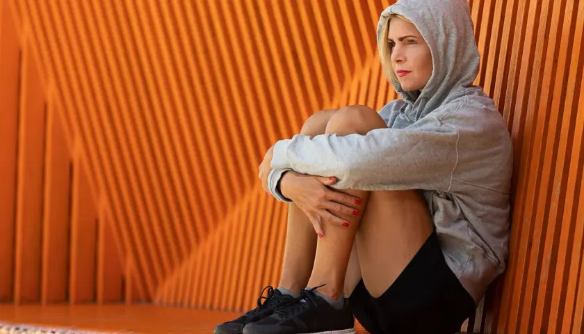 En kvinna i grå hoodie sitter hopkrupen mot en orange vägg, en bild av mental styrka trots en stunds eftertanke.