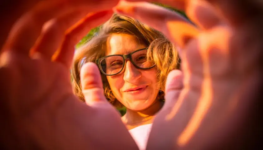Ragazza con occhiali, sorride attraverso le mani che creano una cornice. Un'esperienza emozionale correttiva che illumina il volto.