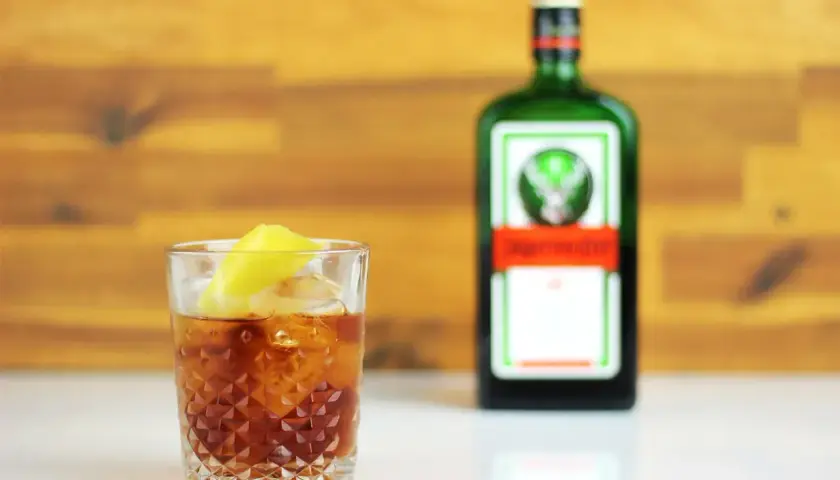Szklanka z drinkiem i cytryną obok butelki Jägermeistera na drewnianym tle.
