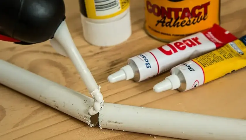 Pistolet z klejem aplikuje masę na pękniętą białą rurkę. Obok leżą tubki kleju, pokazujące, czym skleić plastik.