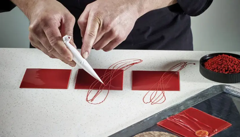 Mains d'un pâtissier créant un décor royal chocolat sur des carrés rouges brillants.