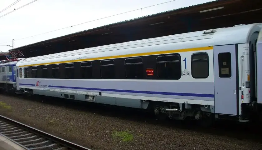 Biało-niebieski wagon PKP Intercity z żółtym pasem, numerem "1" i oknami. To wagon bez przedziałów, idealny na podróż.
