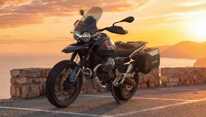 Moto Guzzi V85 TT lista para la aventura al atardecer, con su diseño robusto y maletas laterales, frente a un mar sereno y montañas doradas.