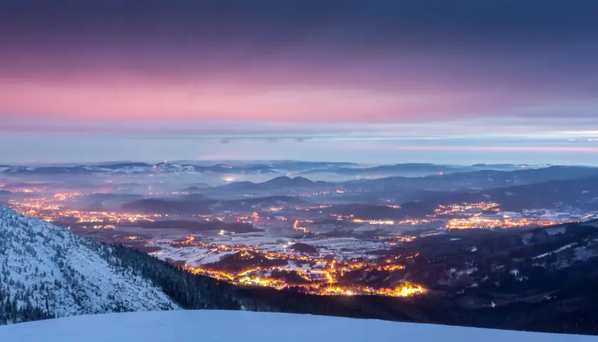 Zachód słońca nad Karkonoszami. Rozświetlone miasto w dolinie, ośnieżone zbocza i zimowe szlaki tworzą magiczny krajobraz.