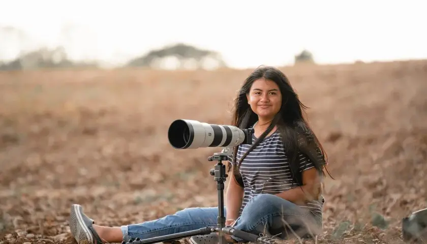 Młoda kobieta z długimi włosami, w pasiastym T-shircie i jeansach, siedzi na ziemi w polu z profesjonalnym aparatem na statywie. To jej fotografia lifestylowa.