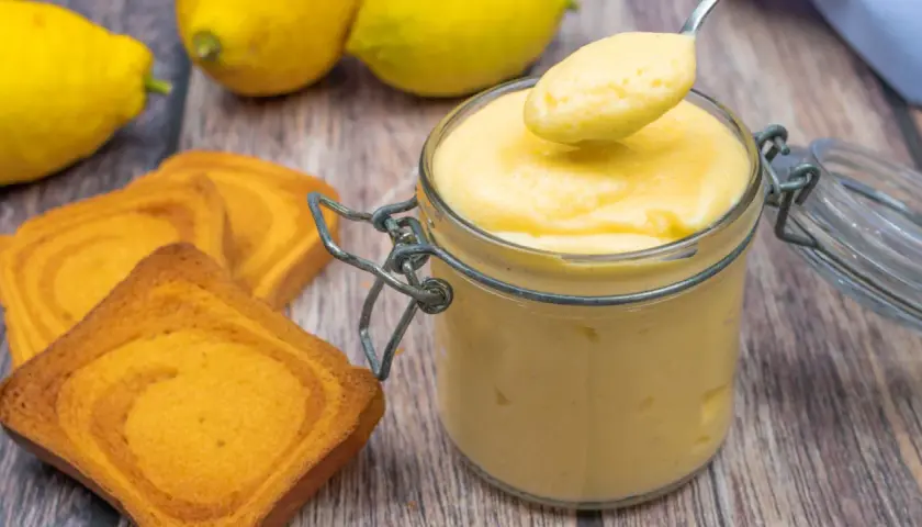Vaso di vetro pieno di cremoso lemon curd, pronto per essere gustato con biscotti e limoni freschi.