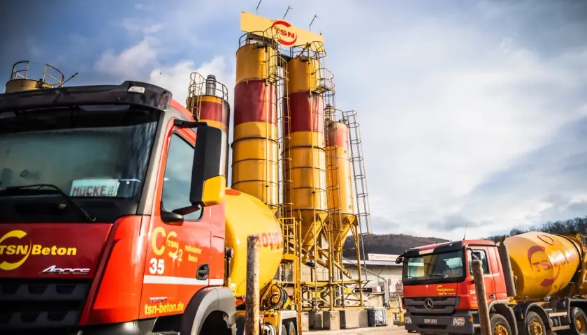 Zwei Betonmischer stehen vor hohen Silos einer Betonmischanlage. Die hgt tragschicht ist bereit für den Transport.