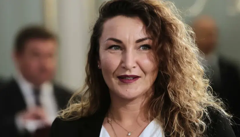 Monika Pawłowska z kręconymi włosami, w białej bluzce i czarnej marynarce, uśmiecha się.