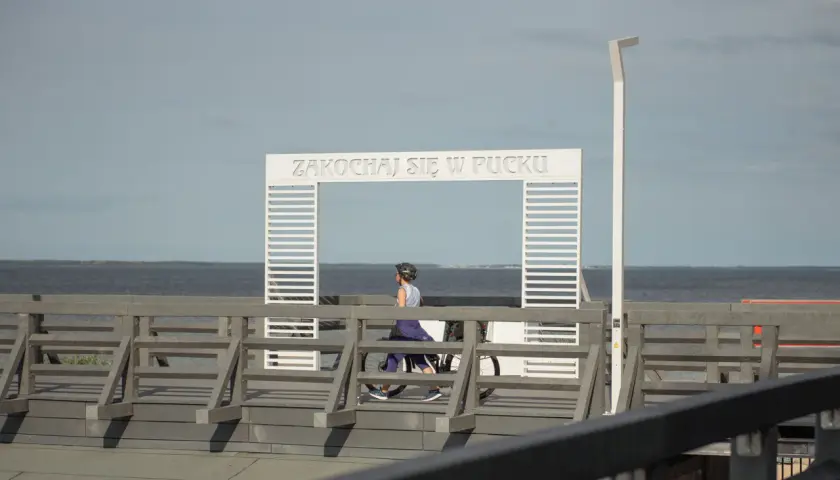 Kobieta na rowerze przejeżdża przez bramę z napisem "Zakochać się w Pucku". Widok na morze z molo w puckim porcie.