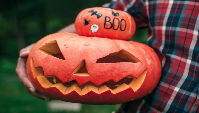 Zucca intagliata con faccia spaventosa e una più piccola sopra con simboli di Halloween in inglese: pipistrello, ragno e la scritta "BOO".
