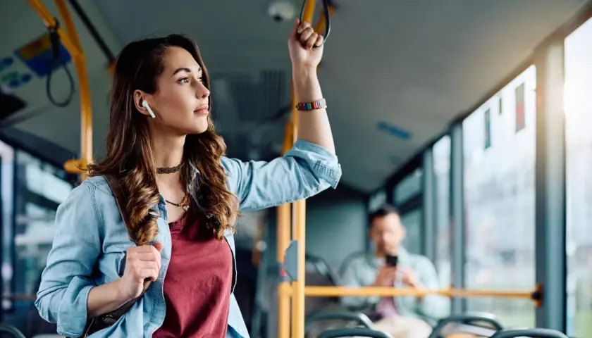 Młoda kobieta w słuchawkach bezprzewodowych trzyma się poręczy w autobusie, słuchając muzyki.