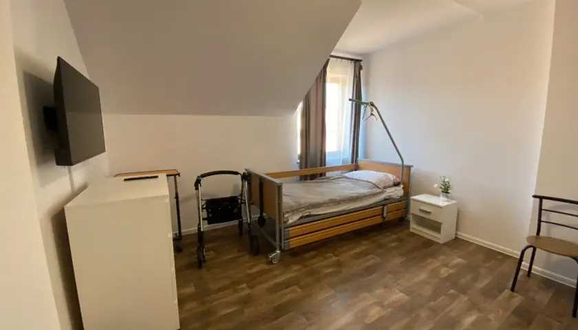 Pokój w domu seniora z łóżkiem, chodzikiem, szafką i telewizorem.