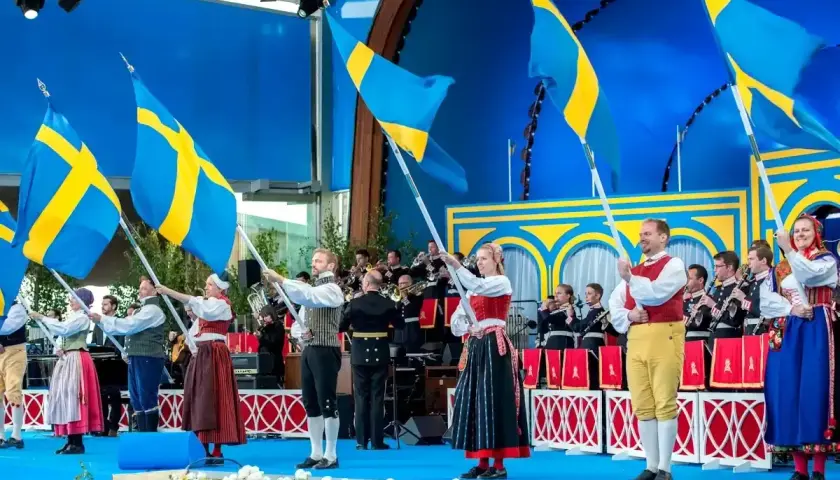 Människor i folkdräkter viftar svenska flaggor under firandet av 6 juni i Stockholm. En orkester spelar i bakgrunden.