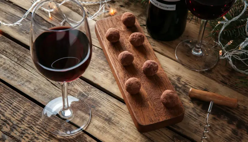 Ein Glas Rotwein und Schokoladentrüffel auf einem Holzbrett, daneben eine Flasche Wein und Lichterketten. Perfekt für einen gemütlichen Abend mit Wein und Schokolade.