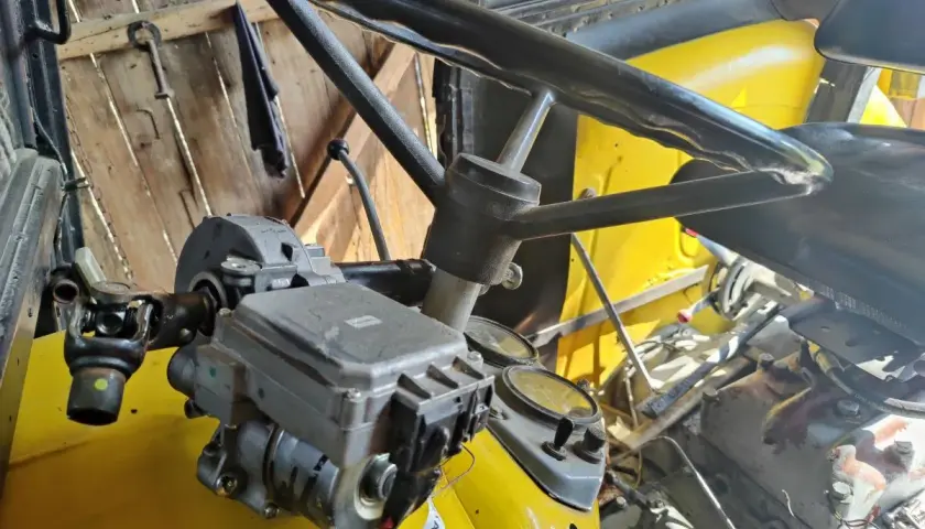 Mechanizm wspomagania kierownicy w żółtym ciągniku. Widoczna kolumna kierownicy, wał napędowy i elementy hydrauliczne.