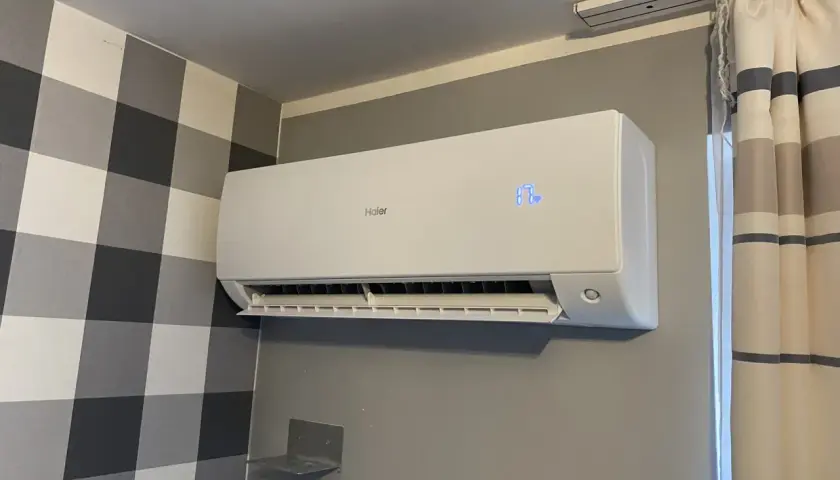 Biała jednostka klimatyzacyjna Haier zamontowana na szarej ścianie, obok kratowanej tapety i zasłon.
