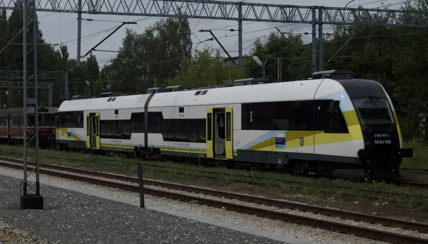Nowy pociąg SA133-008, typu Pesa Elf, z charakterystycznym malowaniem w barwach żółto-biało-niebieskich, stoi na torach. Obok widoczny starszy wagon.