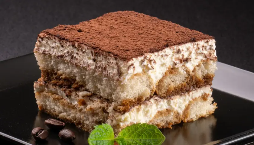 Kawałek tiramisu posypany kakao, z listkami mięty i ziarnami kawy na czarnym talerzu.