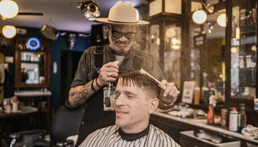 W salonie fryzjerskim "Klasyczna Maszynka" barber w kapeluszu spryskuje wodą włosy klienta przed strzyżeniem.
