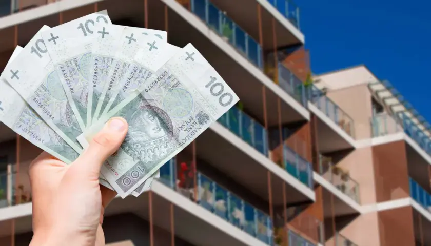 Dłoń trzymająca polskie banknoty 100 zł na tle nowoczesnego budynku mieszkalnego.