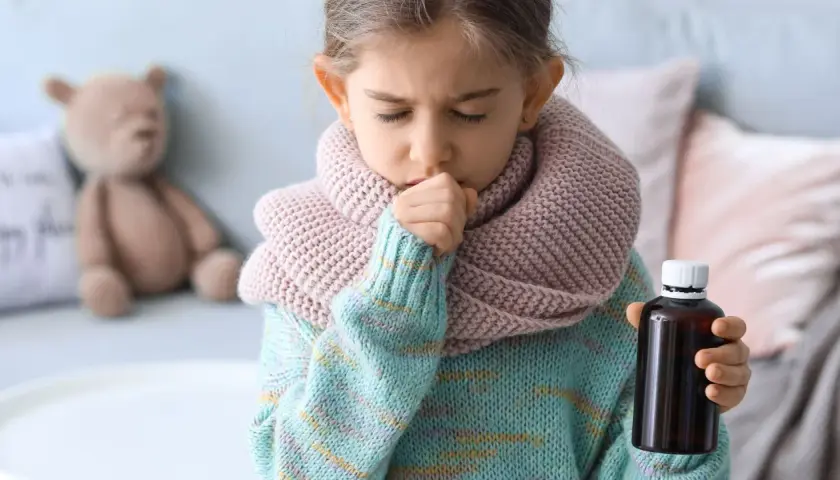 Niña de 3 a 4 años con tos, envuelta en bufanda y suéter, sosteniendo un frasco de medicina.