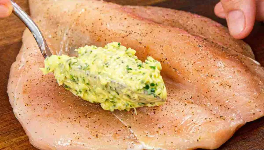 Un cucchiaio di burro alle erbe viene spalmato su un petto di pollo crudo, pronto per essere cucinato.