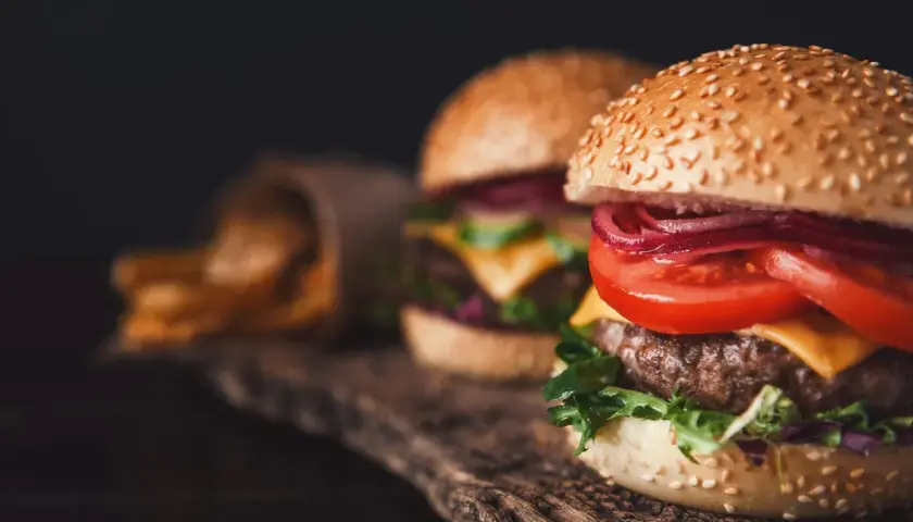 Dwa soczyste smashburgery z serem, pomidorem i czerwoną cebulą na drewnianej desce. Obok kubełek z frytkami.