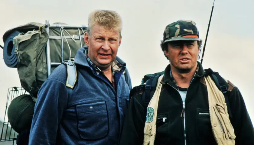 Two men in outdoor gear, one with a large backpack, stand against a pale sky.