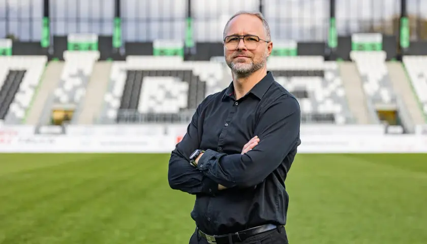 Rafał Smalec, w czarnej koszuli, stoi na tle stadionu piłkarskiego.