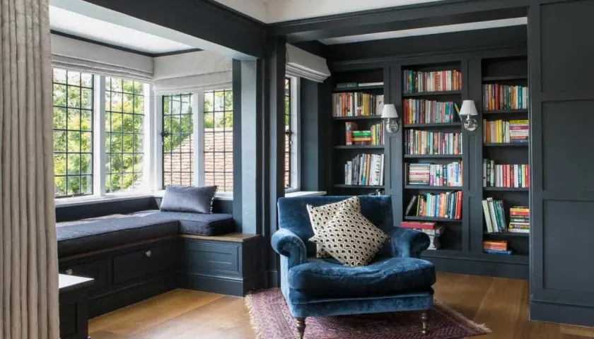 A plush blue armchair sits in a corner cozy reading nook, beside a window seat and floor-to-ceiling bookshelves filled with colorful books.