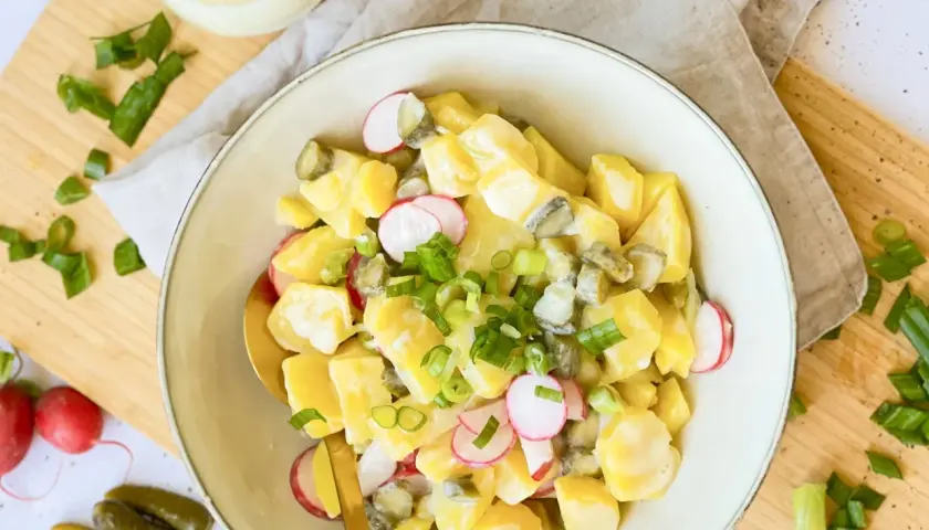Ein cremiger veganer Kartoffelsalat mit Mayo, Radieschen und Gurken, garniert mit Frühlingszwiebeln.