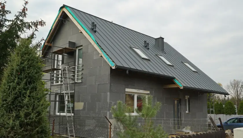 Rozbudowa starego domu w toku. Szary, nowoczesny budynek z metalowym dachem i oknami dachowymi, otoczony rusztowaniami.