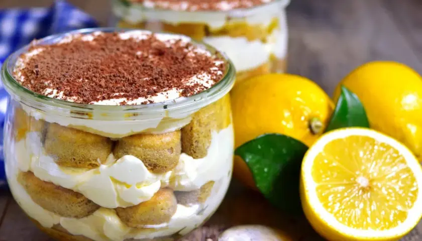 Pyszne tiramisu cytrynowe w słoiczku, przełożone biszkoptami i kremem, posypane czekoladą. Obok świeże cytryny.