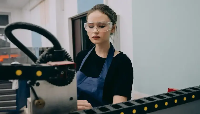 Młody pracownik produkcji w okularach ochronnych i fartuchu pracuje przy maszynie.