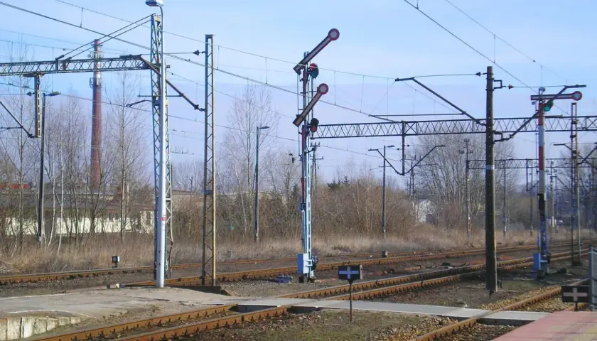 Sygnały na semaforach kolejowych wskazują drogę pociągom. W tle widać komin i drzewa.