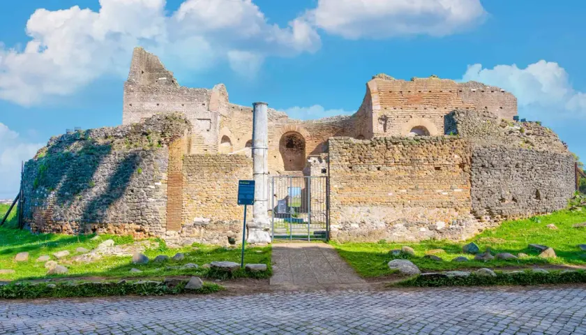 Ruines antiques sur l'Appia Antica à Rome, sous un ciel bleu parsemé de nuages blancs.