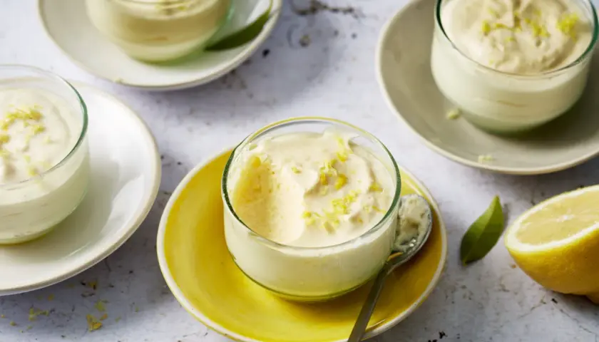 Plusieurs verrines de mousse au citron, garnies de zestes, sur des assiettes claires et jaunes. Un citron coupé est visible à droite.