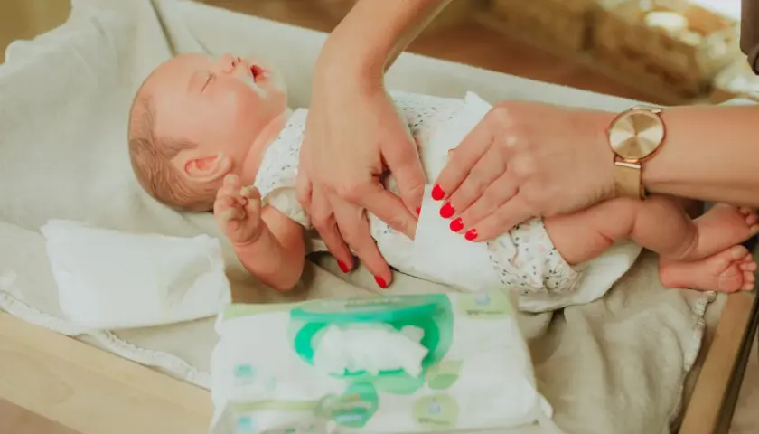 Delikatne dłonie z czerwonymi paznokciami pokazują, jak przewijać noworodka, używając chusteczek nawilżanych.