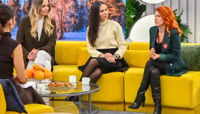 Trzy kobiety siedzą na żółtej kanapie. Jedna z nich, z rudymi włosami, ma na klapie marynarki przypinkę w kształcie serca, jakby dziewczyna wysyła serduszka.