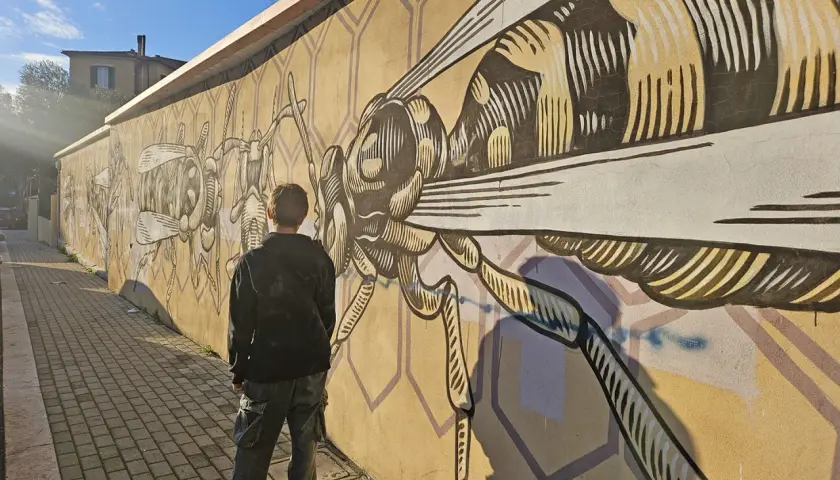 Ragazzo ammira un murale di insetti stilizzati a Quadraro, Roma. La parete gialla è decorata con un'ape gigante e altre creature.