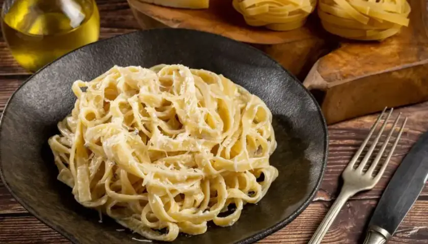 Talerz pełen tagliatelle z kremowym, oryginalnym sosem alfredo, obok butelka oliwy, ser i surowy makaron.