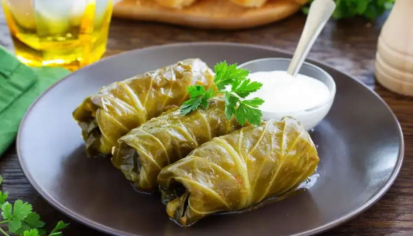 Tre involtini di cavolo cappuccio, guarniti con prezzemolo, serviti con una ciotolina di salsa bianca e un bicchiere di bevanda dorata.