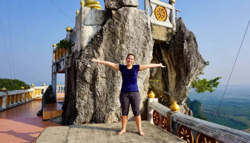 Frau steht vor einem Felsen, der wie ein Tigerhöhlentempel aussieht. Stufen führen zum Tempel.