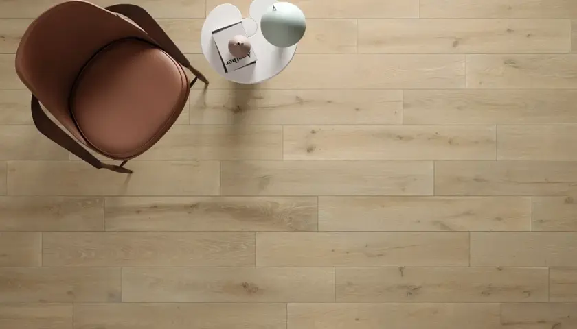 Un tavolino bianco con libro e lampada, accanto a una poltrona marrone, su un pavimento in legno con schemi posa a correre sfalsata.