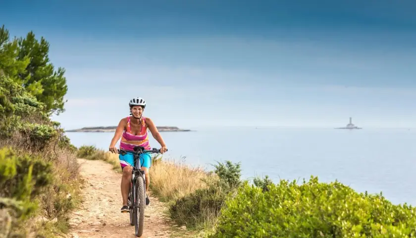 Woman enjoys cycling tours in Slovenia, biking along a coastal path with the sea and a distant lighthouse.