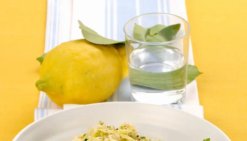 Un piatto di riso zucchine e limone, con un limone fresco e un bicchiere d'acqua.